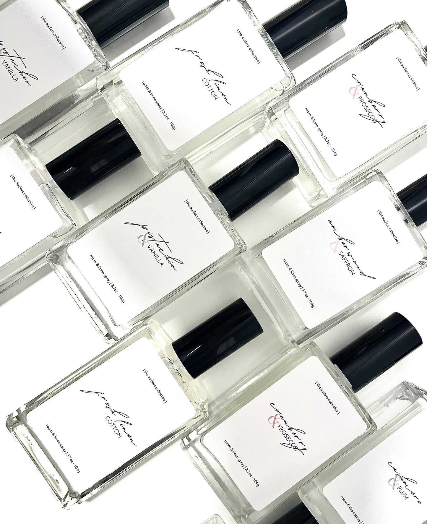 A collection of room and linen spray glass bottles with black caps, each labeled differently with various scent names.