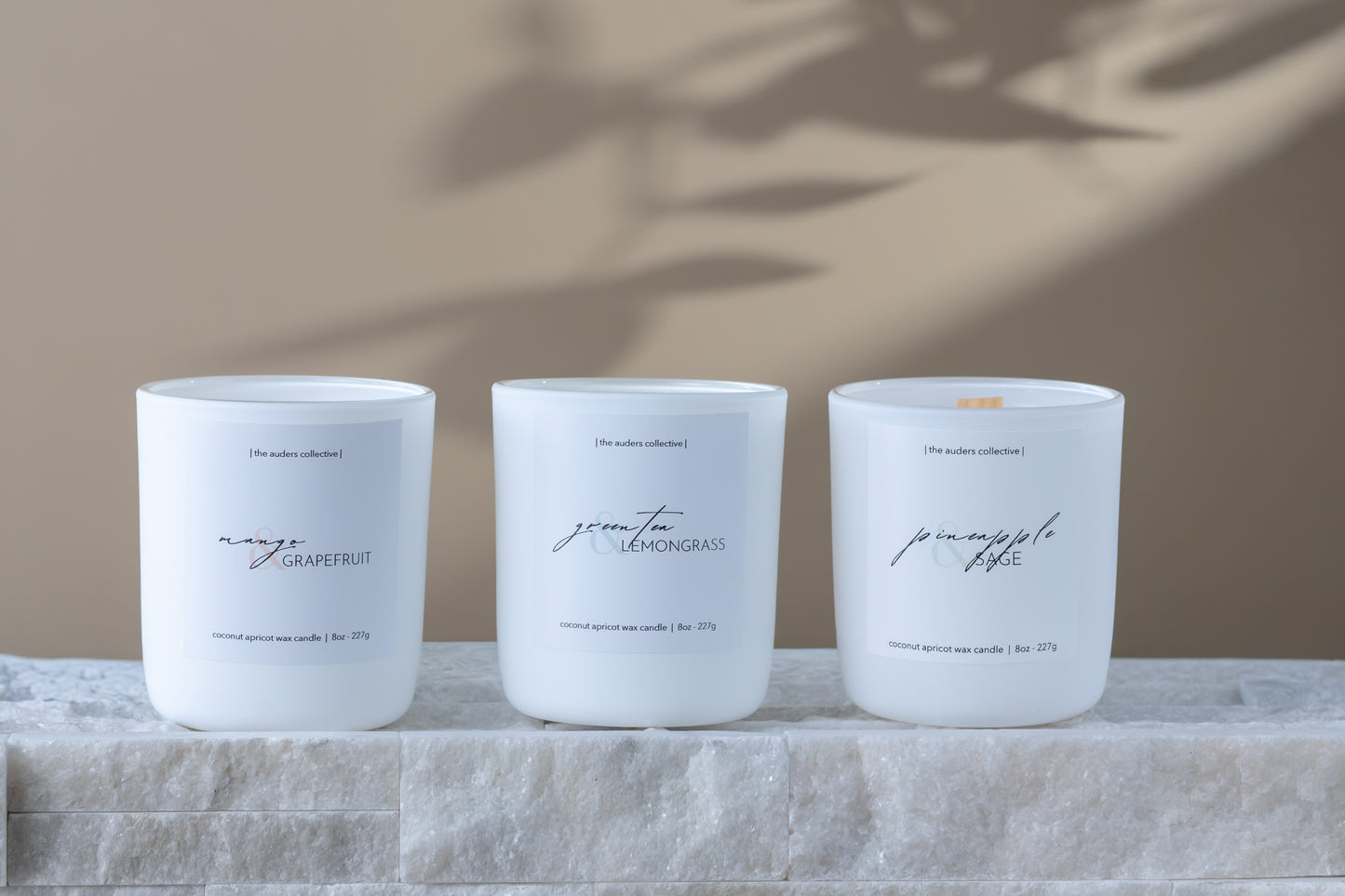 three spring candles on rock tile ledge, white matte jars with minimalist labels - scents are mango and grapefruit, green tea and lemongrass and pineapple and sage.
