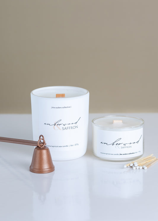 A white candle with a wooden wick, labeled 'Amberwood & Saffron', alongside a copper candle snuffer and matches, on a light background.
