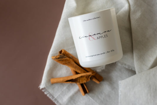 A white candle with the scent cinnamon and apples laying on its side next to white fabric and cinnamon sticks 