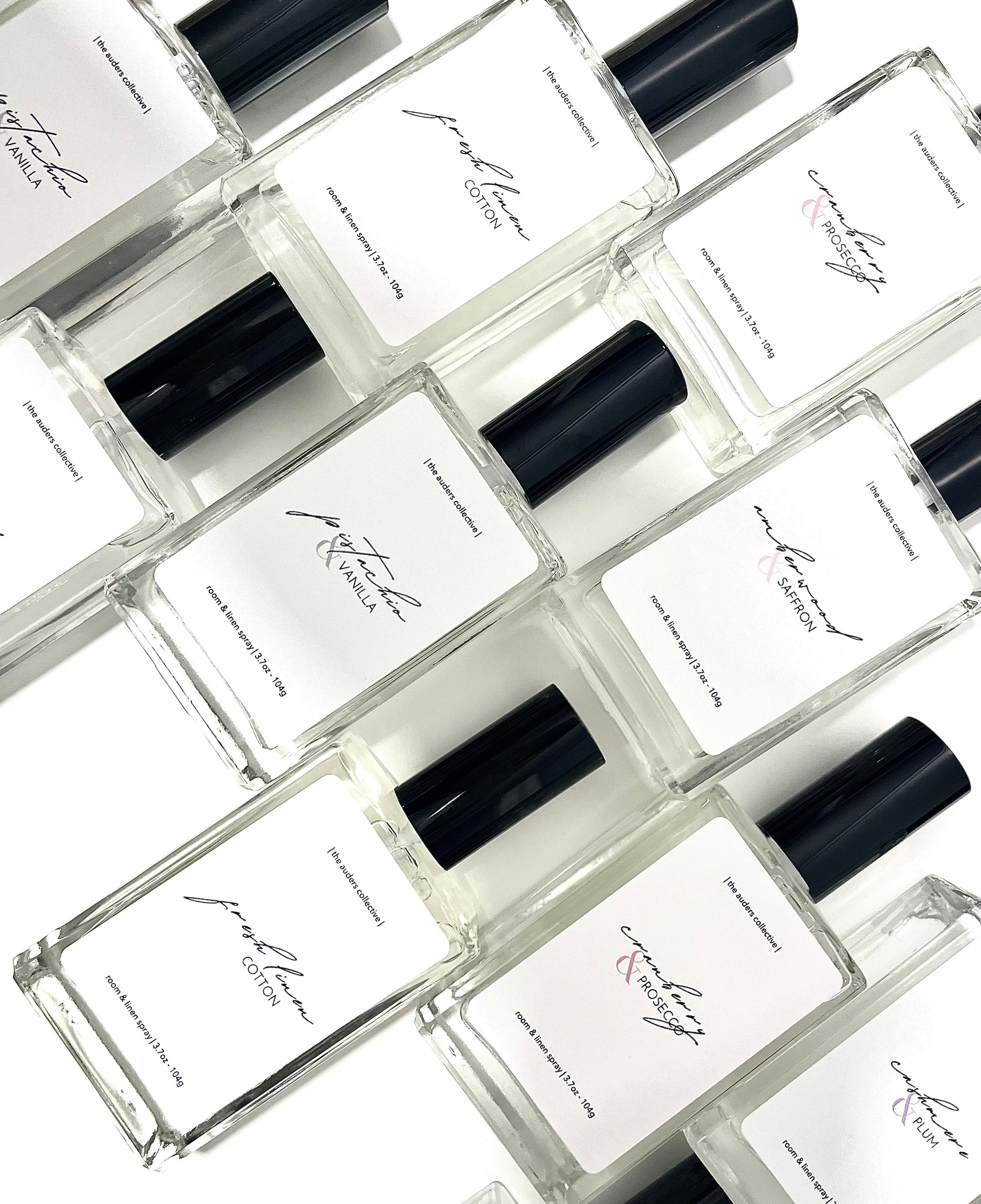 A collection of room and linen spray glass bottles with black caps, each labeled differently with various scent names.