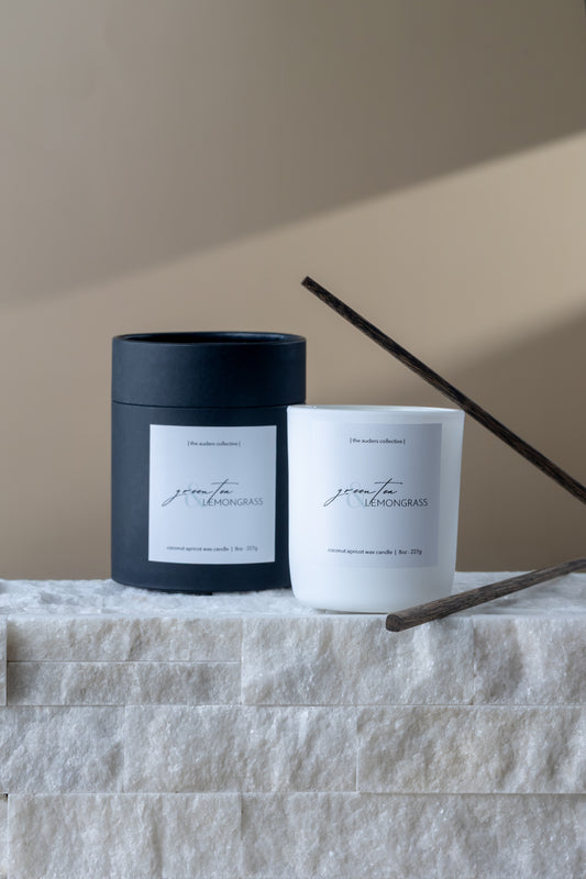 Minimalist green tea and lemongrass scented candle in white glass jar with black packaging on textured stone ledge with soft natural lighting
