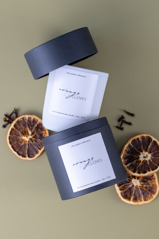 A white candle in between a black candle tube package labeled 'Orange & Cloves' with dried orange slices and cloves on a green background.