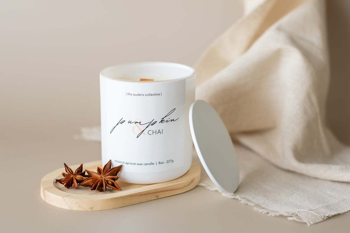 A white candle with 'Pumpkin & Chai' scent written on it, placed on a wooden plate, with a beige fabric and an anise star next to it.