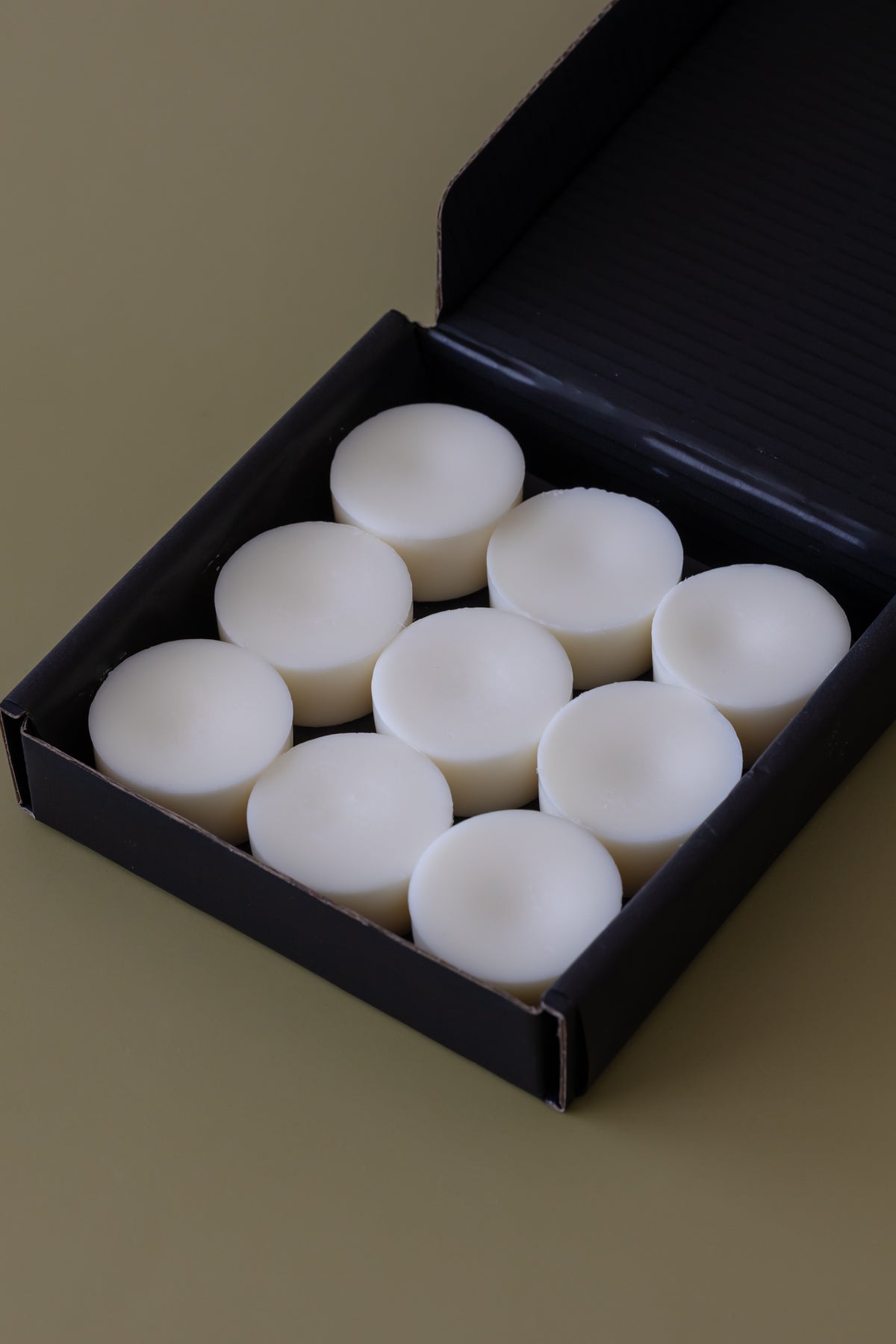 A black gift box containing nine small white wax melts, photographed on a plain warm beige background.