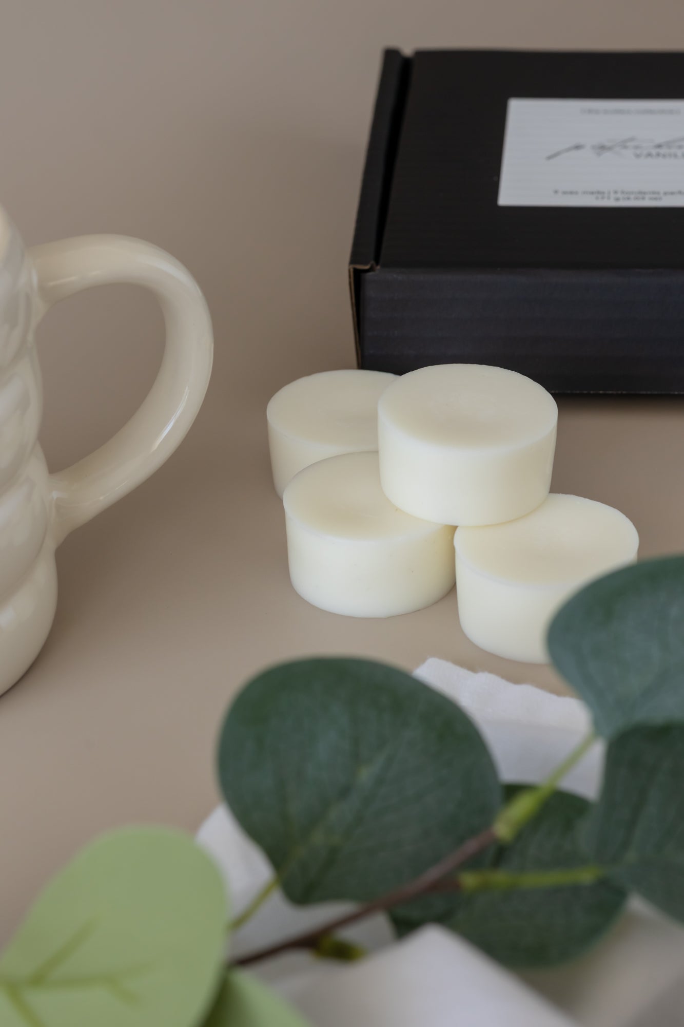 Four small white wax melts arranged on a beige surface next to a black gift box, with a cream ceramic mug partially visible on the left and green leaves in the foreground.